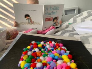 dziewczynka siedzi na łóżku, trzyma książkę "A królik słuchał. na łóżku leży jej praca plastyczna z naklejonymi na czarnej kartce kolorowymi pamponami