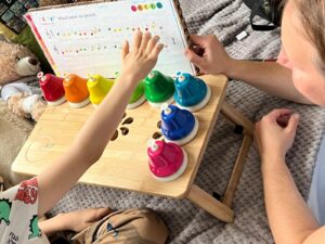 drewniany stolik na łóżku szpitalnym, na nim kolorowe dzwonki i książka z nutami melodii wlazł kotek na płotek. dziecięca ręka sięga do zielonego dzwonka