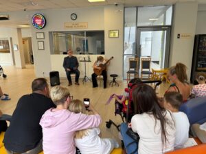 dziewczynka ze szkoły muzycznej grająca na gitarze w holu szpitala. na widowni rodzice i uczniowie