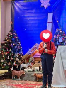 Obraz przedstawia chłopca śpiewającego kolędy. W tle jest choinka oraz żłóbek.